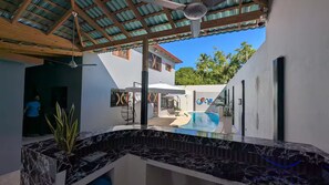Ein moderner Poolbereich im Freien mit Marmorplatte, überdachter Sitzgelegenheit und Blick auf ein Haus im Hintergrund.
