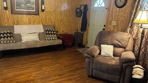 Un salon chaleureux avec un plancher en bois, un canapé, un fauteuil inclinable, une valise rouge et une porte avec une fenêtre.
