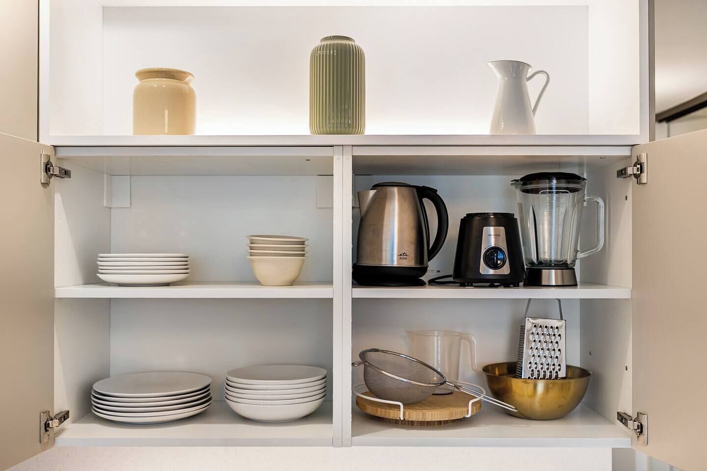A kitchen cabinet with shelves containing plates, bowls, a kettle, a blender, a strainer, and a grater.
