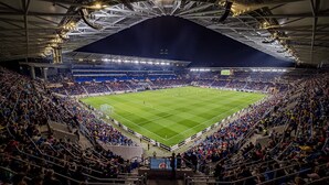 Een groot overdekt stadion met meerdere rijen zitplaatsen, een veld in het midden en verlichting erboven.