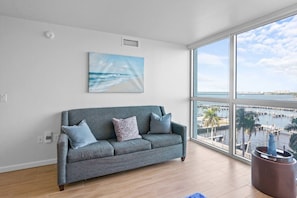 Ein modernes Wohnzimmer mit einem grauen Sofa, einem Strandbild und einem großen Fenster mit Blick auf eine Marina.