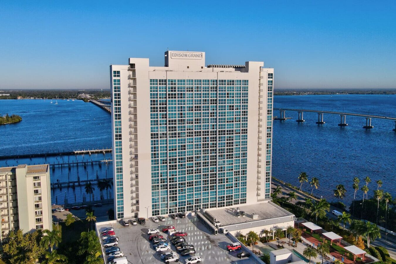Un grand hôtel, avec une façade en verre imposante, un parking et une vue sur une étendue d’eau, avec un pont en arrière-plan.