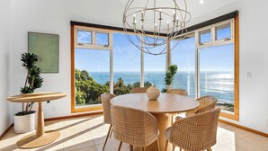 Un comedor con una mesa redonda y sillas de mimbre, ventanas amplias con vistas al océano y un candelabro.
