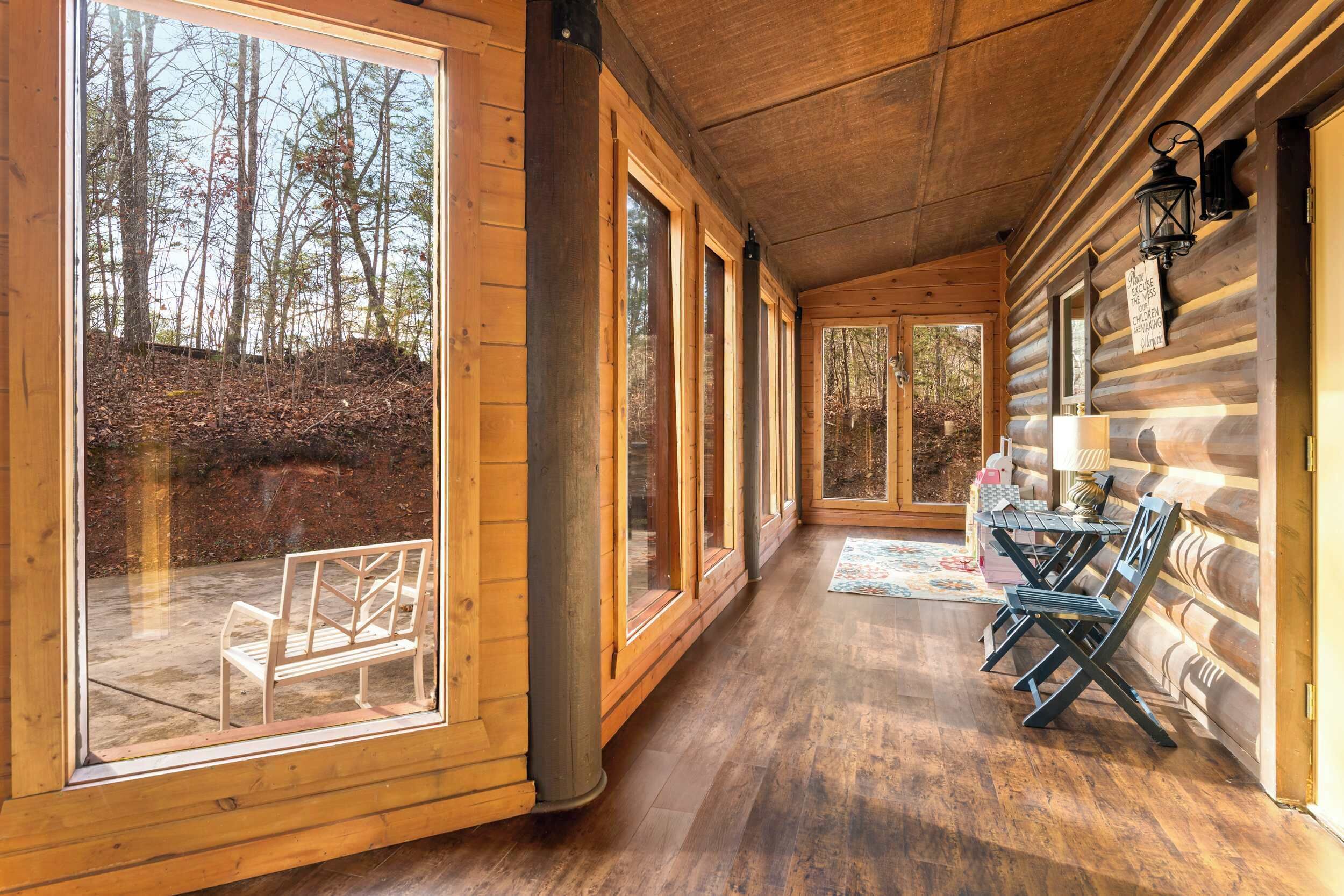 A log cabin interior with large windows, a wooden floor, and a dining area with chairs and a table.