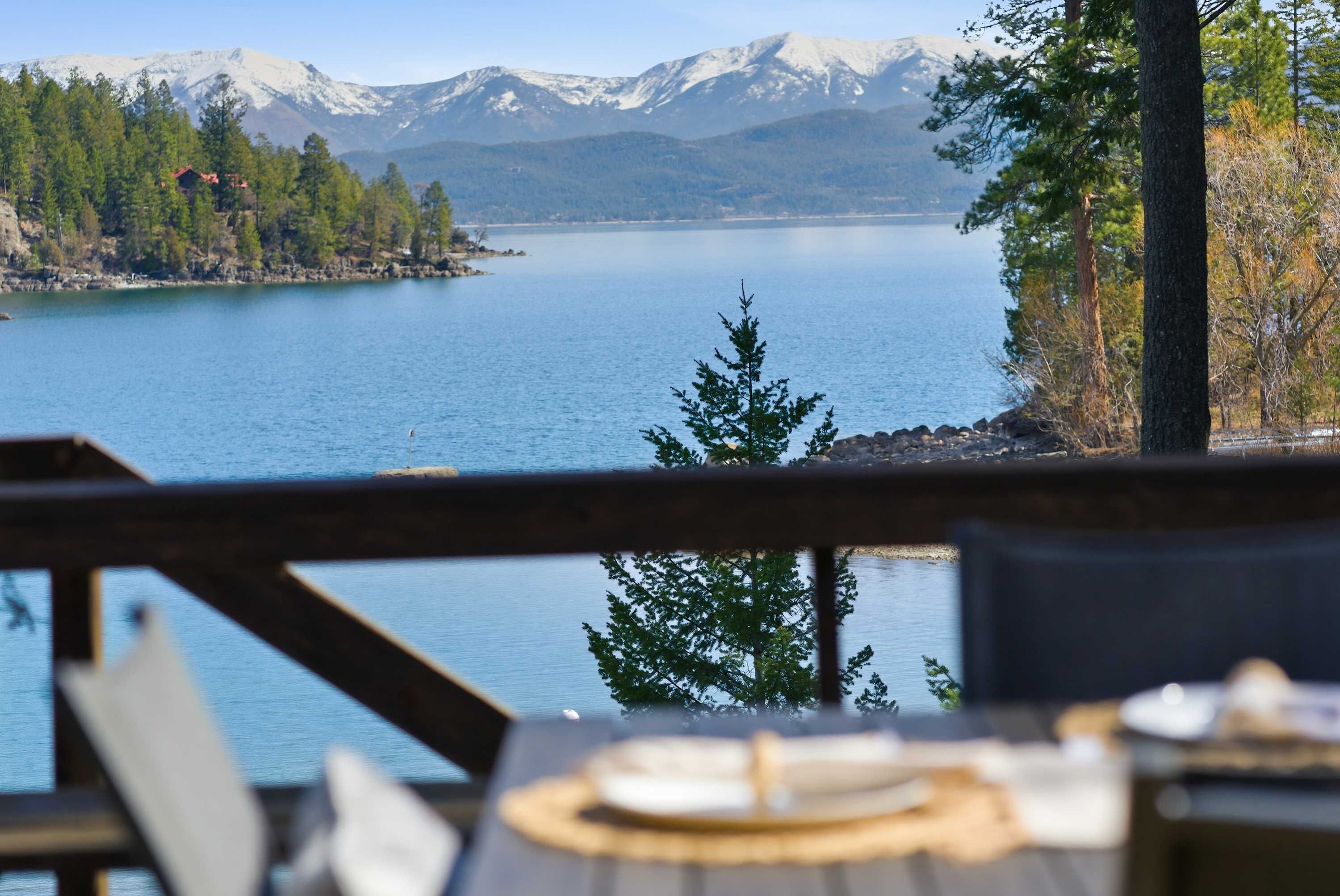 Blick auf einen See mit Bergen im Hintergrund, eine Holzterrasse und ein Tisch mit Unterlage.
