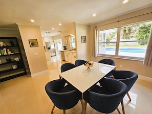 A modern kitchen with white cabinets, a stainless steel refrigerator, and a dining area with a white table and four dark chairs. - Modern Pool Home- 35th Ave (Vero Beach South)