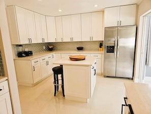 A modern kitchen with white cabinetry, a stainless steel refrigerator, and a central island with a black stool. - Modern Pool Home- 35th Ave (Vero Beach South)
