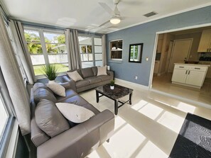 A spacious living room with a large sectional sofa, a coffee table, and a view of the backyard through large windows. - Modern Pool Home- 35th Ave (Vero Beach South)