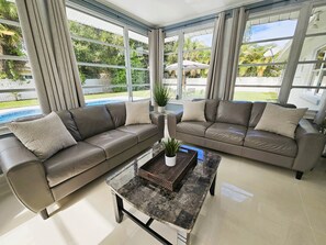 A modern living room with a sectional sofa, a coffee table, and large windows offering a view of a backyard with a pool. - Modern Pool Home- 35th Ave (Vero Beach South)