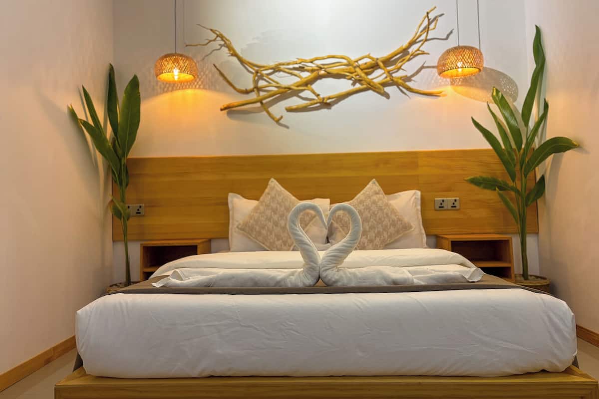 A neatly made bed with white linens and two heart-shaped towels on the pillows. There are two wall-mounted lamps with warm lighting, and a decorative branch on the wall above the bed.
