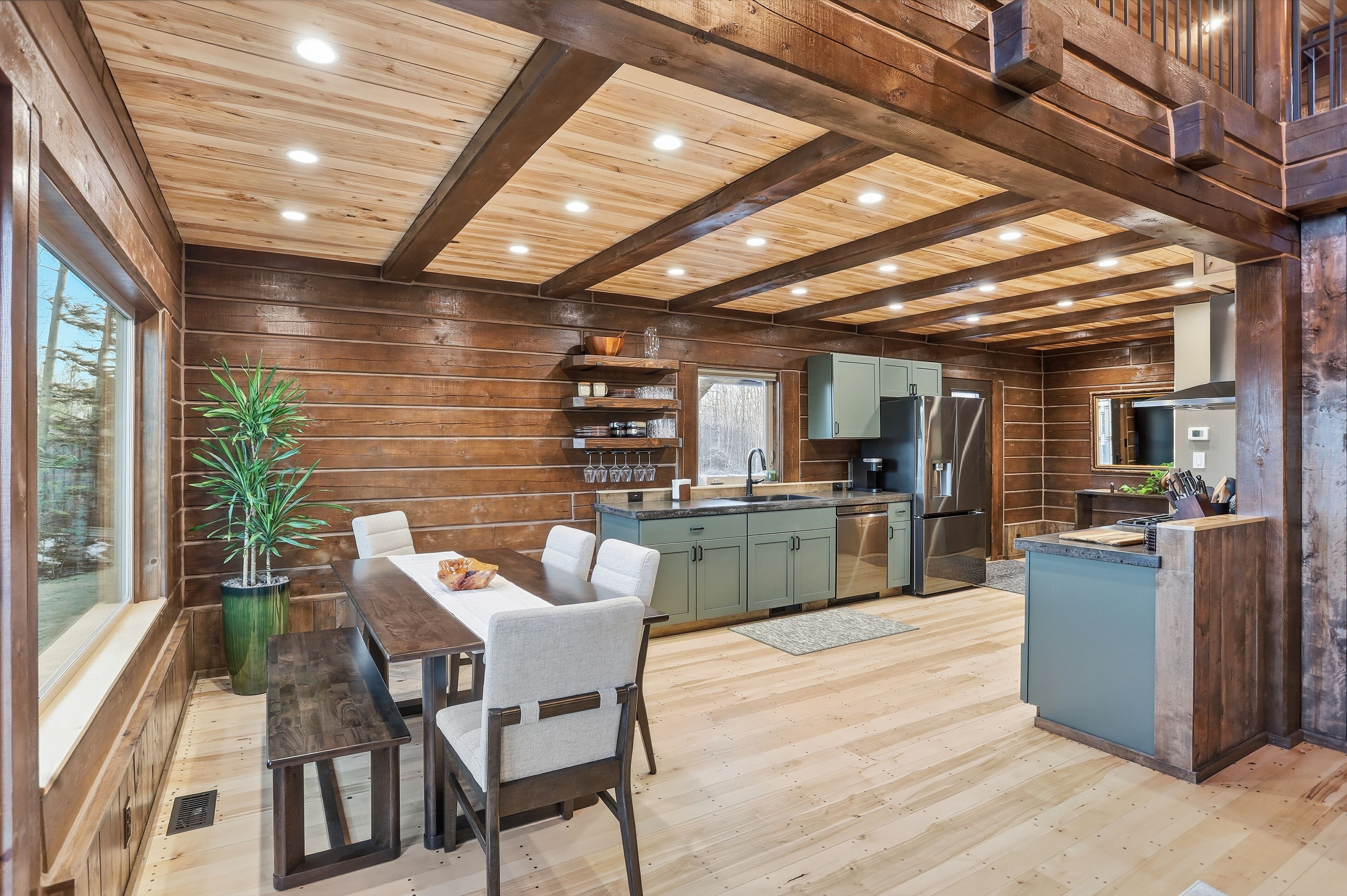 A modern kitchen with wooden beams, a dining area with a table and chairs, and a large window overlooking a forest.