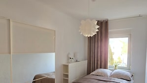 A bedroom with a bed, a large mirror, a bedside table, a window with curtains, and a hanging light fixture.