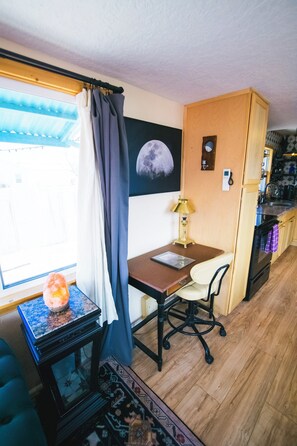 A compact living space with a desk, chair, and a small table with a candle. A window with curtains and a view of the outside is visible.