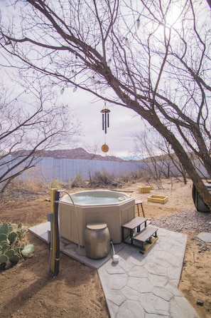 A hot tub with a wooden bench and a table, surrounded by a patio area with a cactus plant and a view of a lake and mountains.