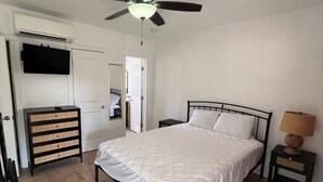 Un dormitorio con cama, mesita de noche, cómoda, ventilador de techo y televisor en la pared.