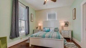 A bedroom with a bed, two bedside tables, a ceiling fan, and a window with curtains. - Surf House Galveston (Galveston)