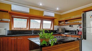 A kitchen with wooden cabinets, a black countertop, a refrigerator, a microwave, and a window with a view of greenery. - Casa Confortável em Angra do Reis Proximo a Praia (Angra dos Reis)