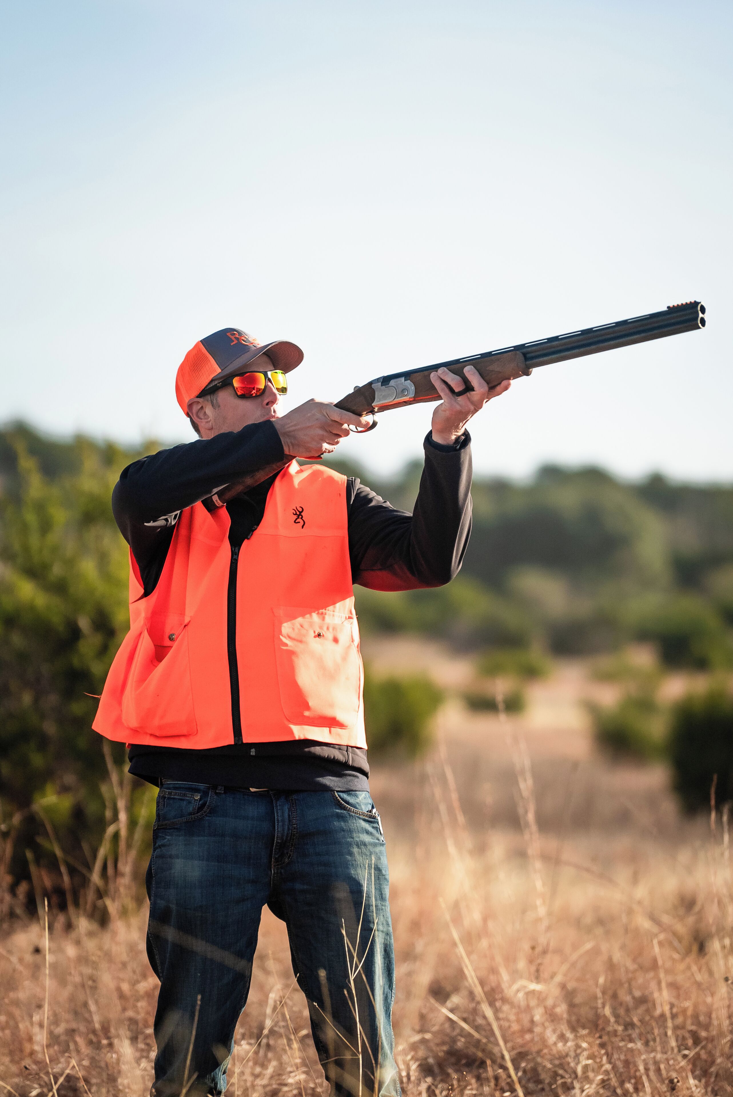 A person in an orange vest aiming a shotgun in an outdoor setting.