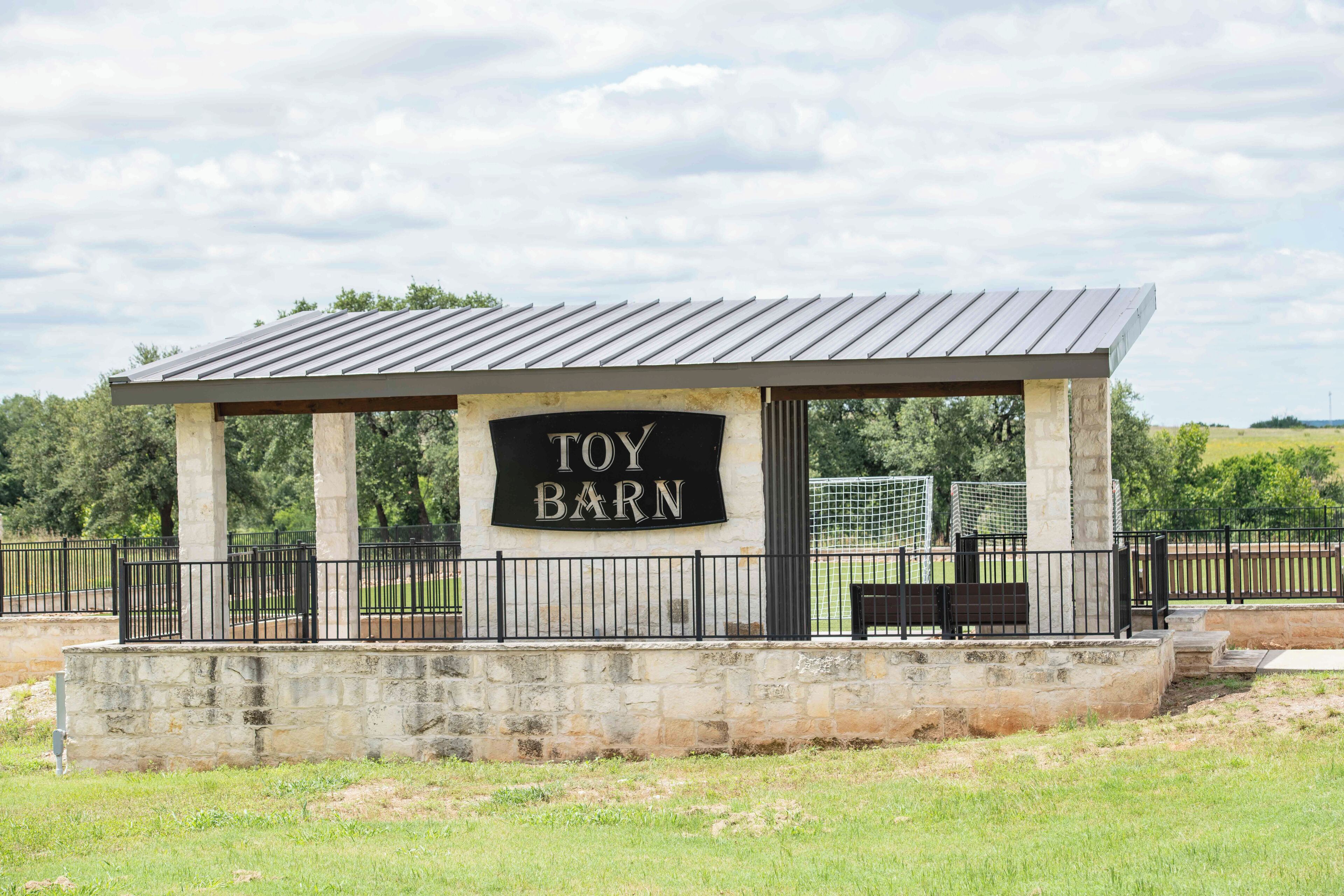 A structure labeled "TOY BARN" with a metal roof and stone pillars, surrounded by a black metal fence.