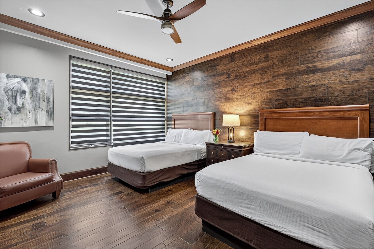 A hotel room with two beds, a ceiling fan, a large window with blinds, a wooden headboard, a nightstand with a lamp, and a painting on the wall.
