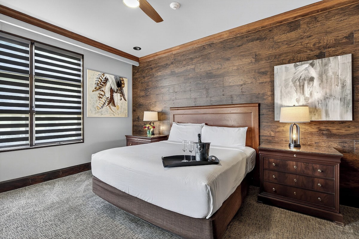 A bedroom with a large bed, wooden headboard, a nightstand with a lamp, and artwork on the walls.
