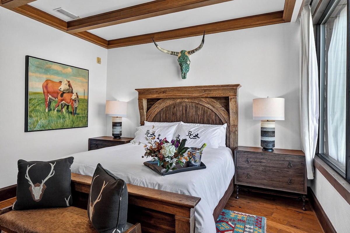 A bedroom with a wooden headboard, a bed with white linens, two bedside tables with lamps, a painting of cows, and a decorative skull on the wall.