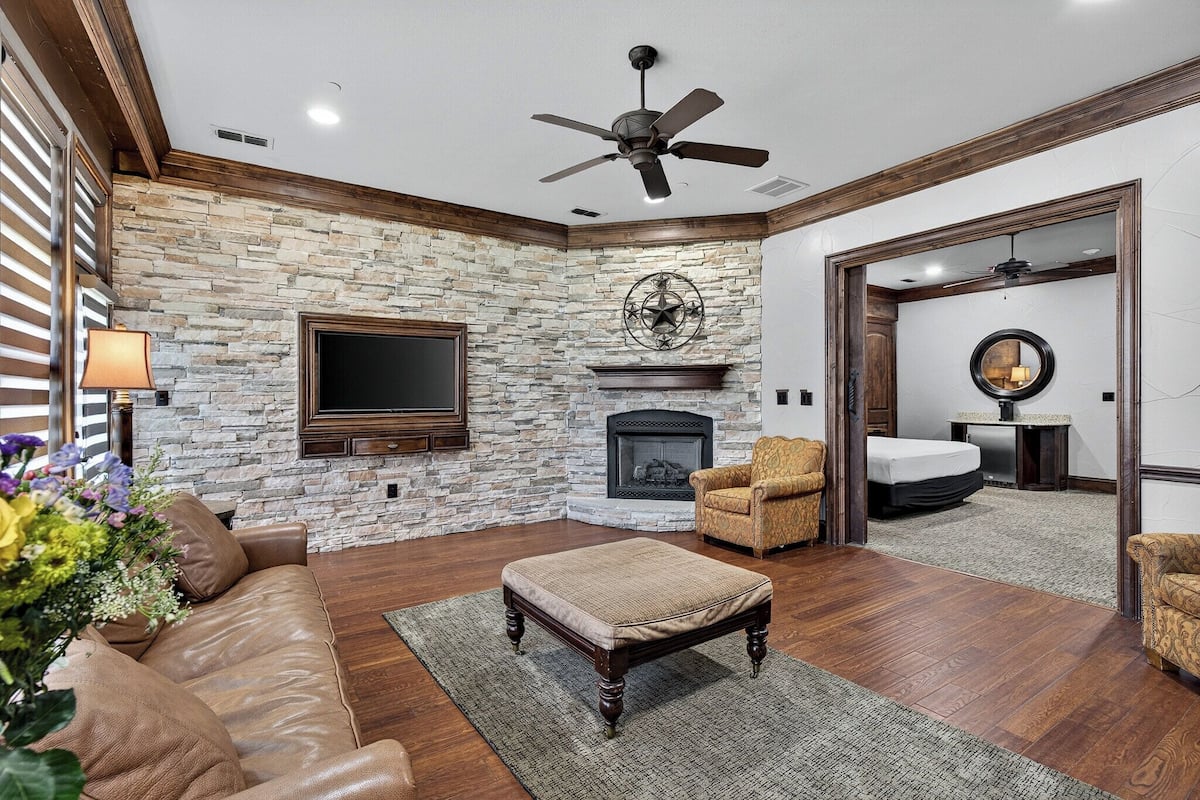 A living room with a stone wall, a fireplace, a ceiling fan, and a TV mounted on the wall.