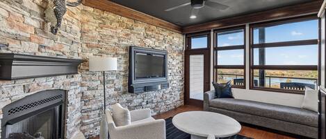 A room with a stone wall, a fireplace, a flat-screen TV, a ceiling fan, and a large window with a view.