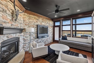 Una habitación con pared de piedra, chimenea, televisor de pantalla plana, ventilador de techo y un ventanal con vistas.