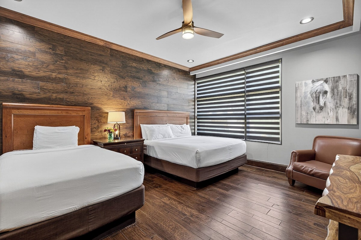 A hotel room with two beds, wooden flooring, a ceiling fan, a window with blinds, and a painting on the wall.