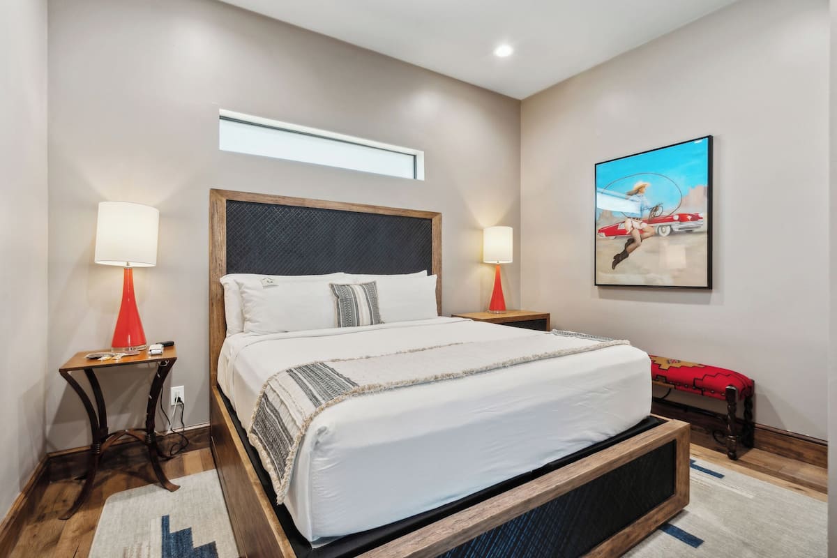 A bedroom with a large bed, two bedside tables with lamps, a window, and a painting on the wall.