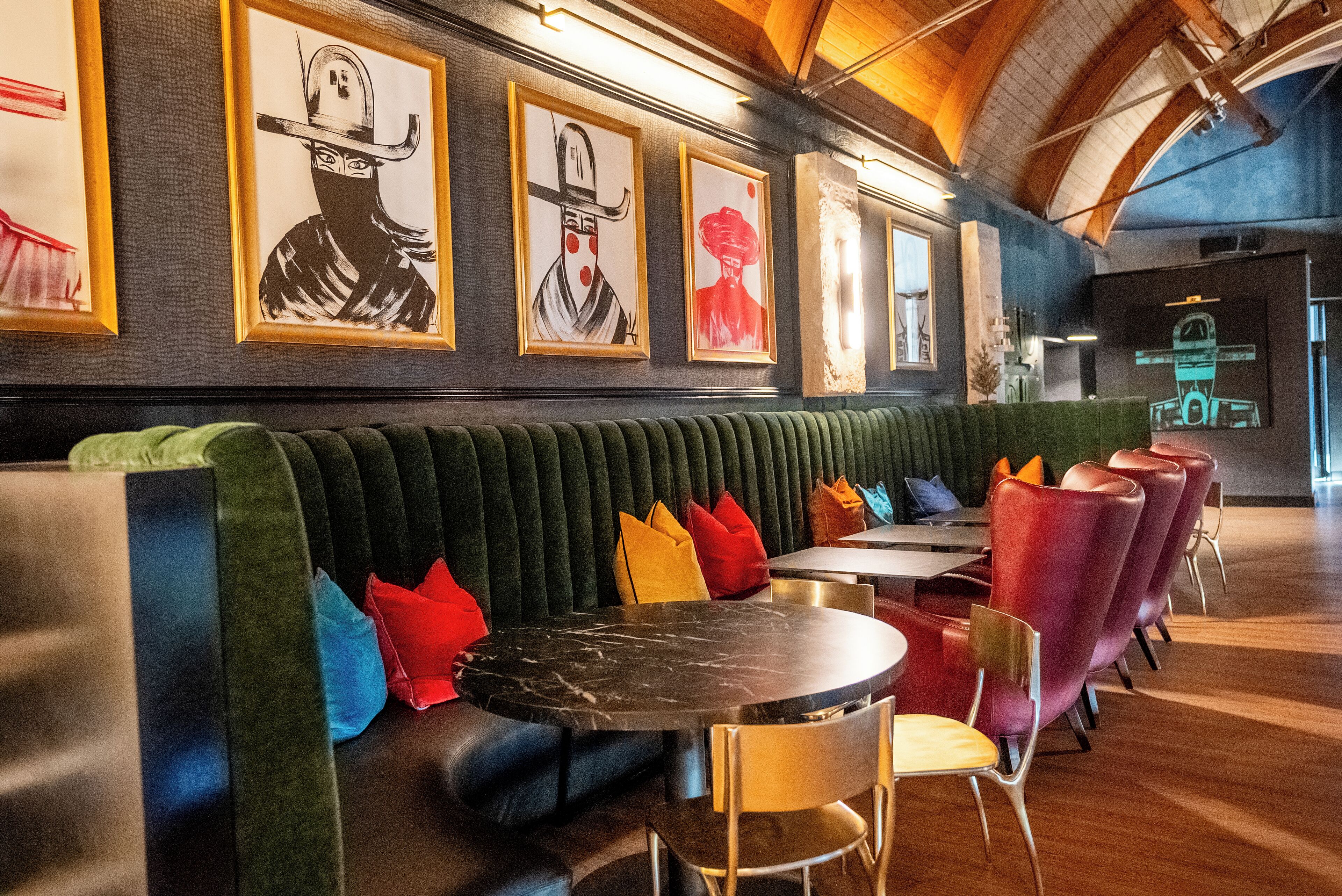 Een modern restaurant met een lange, groene gestoffeerde bank, rode en gele kussens, en een marmeren tafel.