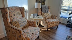 Een zithoek met gestreept fauteuils, een klein rond tafeltje met een lamp, en uitzicht op het omliggende landschap door een raam.