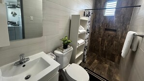 Een badkamer met een witte wastafel, toilet en douchegedeelte. Boven de wastafel hangt een spiegel en op het aanrecht staat een plant.