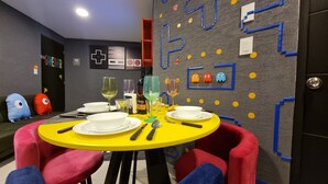 Un área de comedor con una mesa amarilla, sillas rojas y azules, y una pared decorada con patrones geométricos y pequeñas figuras.