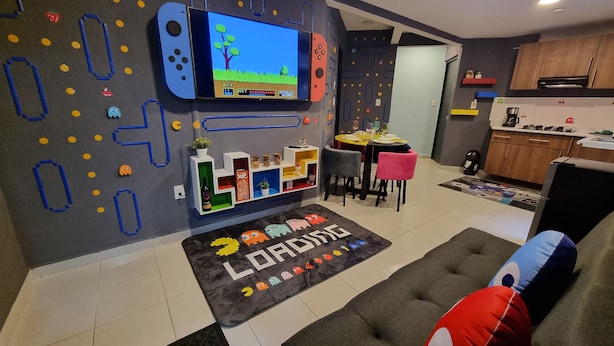 Una sala de estar con una consola de videojuegos, un sofá, una mesa de centro y un comedor con sillas y una mesa.