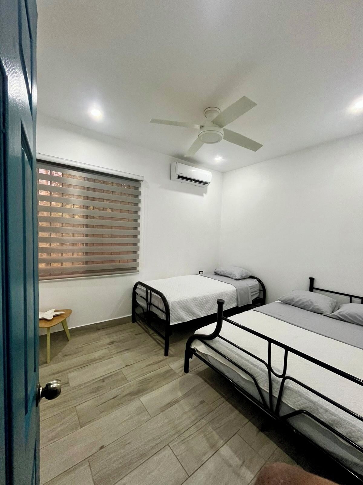 A dormitory room with two beds, a ceiling fan, an air conditioning unit, a window with blinds, and a small table.