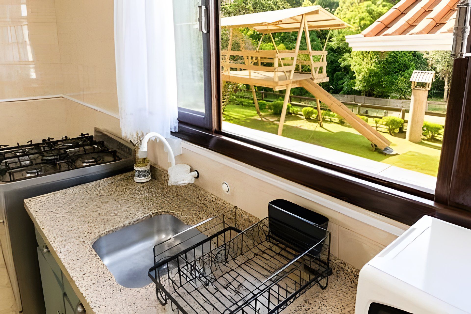 A kitchen with a granite countertop, a stainless steel sink, and a window overlooking a wooden deck with a slide.
