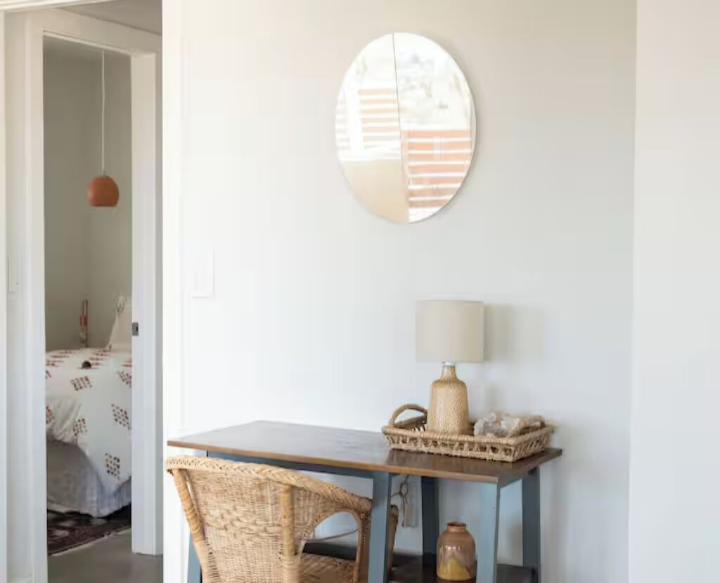 A room with a round mirror on the wall, a small table with a lamp, and wicker furniture.