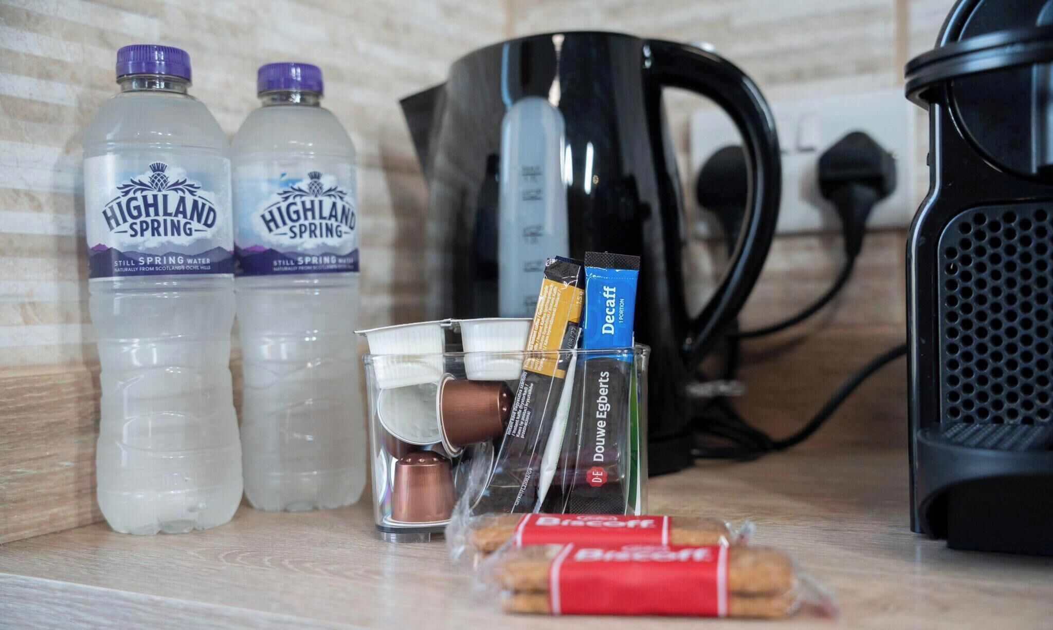 Una encimera de cocina con dos botellas de agua Highland Spring, una cafetera, un hervidor, un vaso y dos paquetes de cigarrillos.