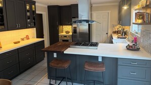 A modern kitchen with dark cabinets, a central island with stools, a stainless steel range hood, and a built-in oven. - 4-bedroom house in perfect Santa Barbara, 5 minutes from the beach! (Santa Barbara)