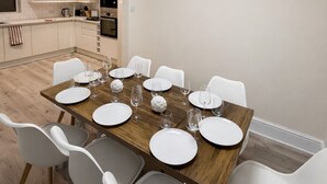 A modern kitchen with white cabinets and a central island, a dining area with a wooden table and white chairs, and a recessed ceiling light. - Beautifully Refurbished Home with 4 Bedrooms (Greenmount)