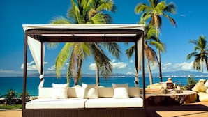 Una cama frente a la playa con dosel, almohadas blancas y vista al océano y palmeras.