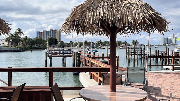 An outdoor seating area with a thatched umbrella, wooden chairs, and a view of the water and buildings in the background. - 216 Panoramic Single Room in a waterfront hotel in Singer Island, FL (Riviera Beach)