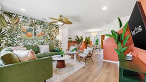 A modern living room with a green sofa, a coffee table, and a dining area with wicker chairs. There is a tropical wallpaper on the wall and a large plant in the corner. - Epic Backyard - Pool - Hot Tub - Outdoor Theatre (Bradenton)