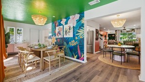 A modern kitchen with wooden cabinets, a dining area with a round table and chairs, and a vibrant tropical mural on the wall. - Epic Backyard - Pool - Hot Tub - Outdoor Theatre (Bradenton)