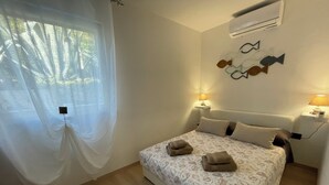A bedroom with a bed, two bedside tables, a wall-mounted air conditioner, and a window with sheer curtains. - Residence4a- Apartman Bianco - u blizini mora, idealan za opuštanje (Ravni)