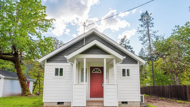 Una casa pequeña de un solo piso con puerta roja, revestimiento blanco y un porche delantero.