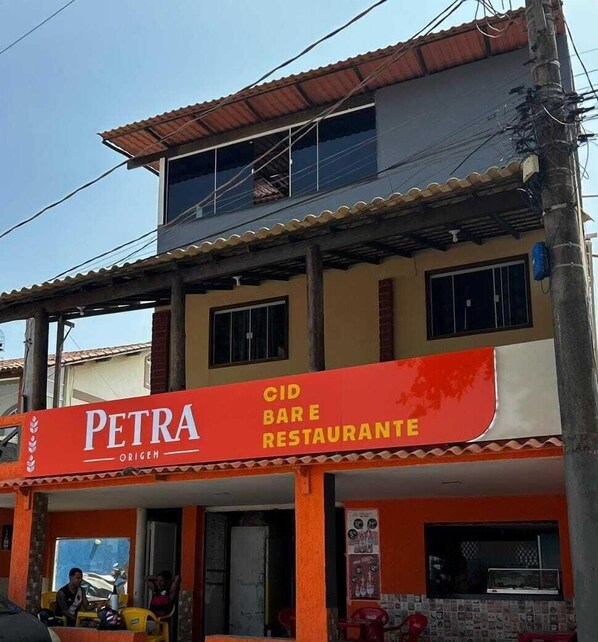A two-story building with a restaurant named "PETRA ORIGEH" on the ground floor. - Suítes Prime (Marataízes)