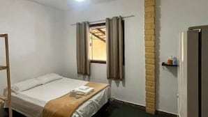 Une petite pièce simple, avec un lit, une fenêtre avec des rideaux, un mur en briques et un réfrigérateur.
